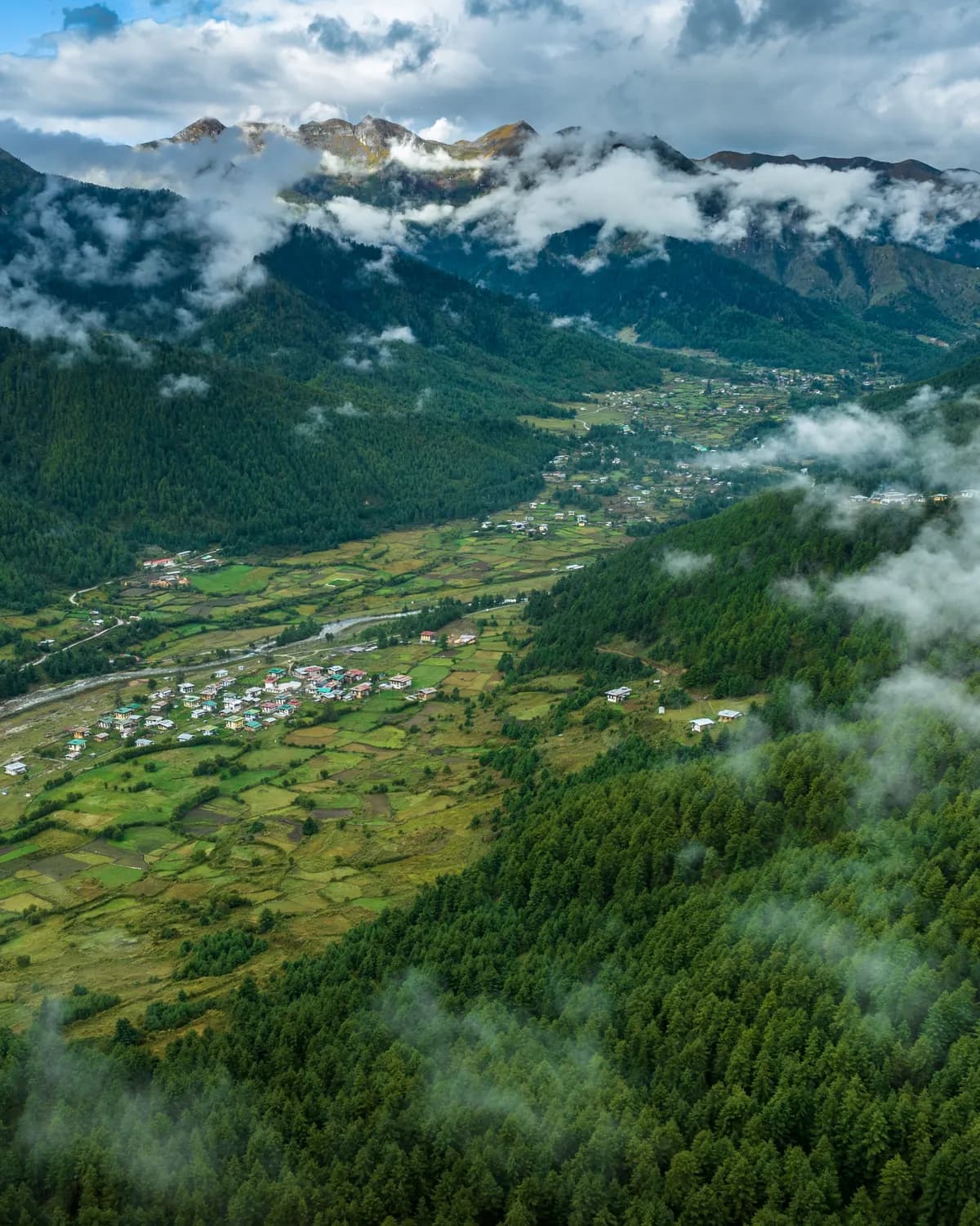 Aerial view of a forested Bhutanese valley with farmland, a village and cloud-wrapped mountains
