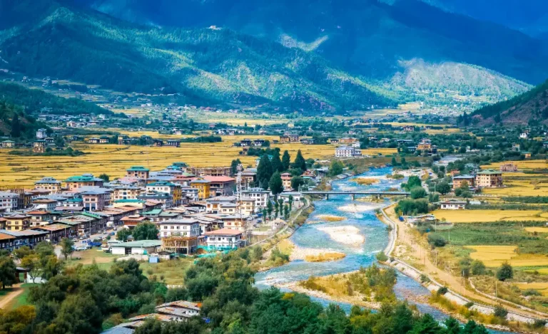 View of Paro town in autumn with colorful trees, traditional Bhutanese buildings, and surrounding Himalayan mountains under a clear blue sky.